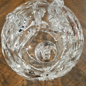 May include: Clear, cut-glass bowl with a round shape and three decorative feet. The bowl features a starburst pattern and intricate detailing. The bowl is made of clear glass and is sitting on a wooden surface.