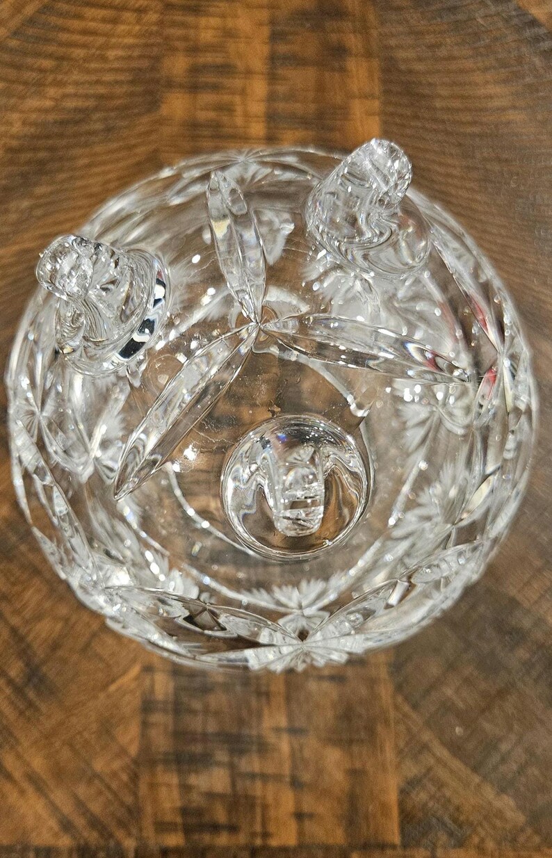May include: Clear, cut-glass bowl with a round shape and three decorative feet. The bowl features a starburst pattern and intricate detailing. The bowl is made of clear glass and is sitting on a wooden surface.