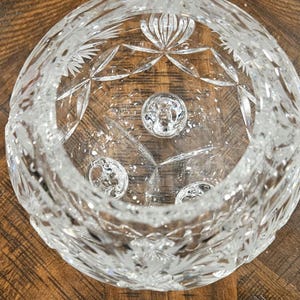May include: A clear, cut-glass bowl with intricate star and floral patterns. The bowl features a circular design with three small, round, decorative elements at the base. The bowl is sitting on a wooden surface.