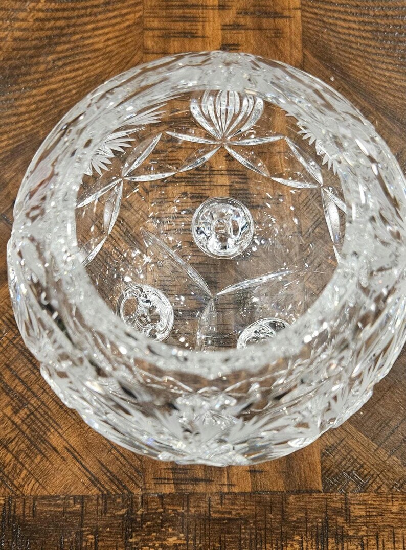 May include: A clear, cut-glass bowl with intricate star and floral patterns. The bowl features a circular design with three small, round, decorative elements at the base. The bowl is sitting on a wooden surface.