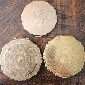 May include: Three vintage gold-toned compact mirrors with ornate designs. One mirror has a detailed floral and crest pattern, while another features a simple, textured surface. The third compact has a repeating circular pattern. All have a scalloped edge.