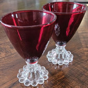 May include: Two ruby red stemmed glasses with clear, bubble-shaped bases. The glasses have a conical bowl shape and are displayed on a wooden surface. The stem and base are clear glass.