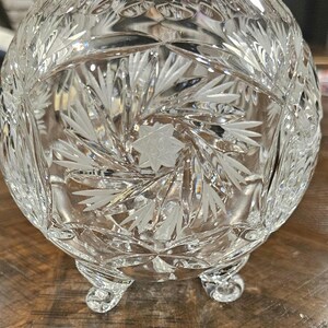 May include: A clear, cut-glass bowl with a rounded shape and three small feet. The bowl features intricate etched designs of starbursts and leaf patterns, creating a sparkling, decorative piece.