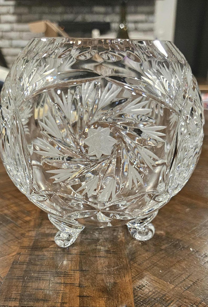 May include: A clear, cut-glass bowl with a rounded shape and three small feet. The bowl features intricate etched designs of starbursts and leaf patterns, creating a sparkling, decorative piece.