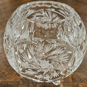 May include: A clear, round crystal bowl with intricate etched floral and geometric patterns. The bowl sits on three small feet and has a wide opening. The cut glass design reflects light, creating a sparkling effect. A decorative piece for display.
