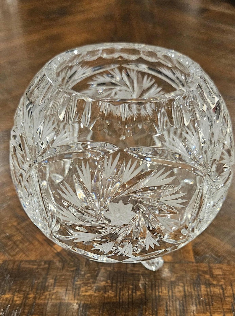 May include: A clear, round crystal bowl with intricate etched floral and geometric patterns. The bowl sits on three small feet and has a wide opening. The cut glass design reflects light, creating a sparkling effect. A decorative piece for display.