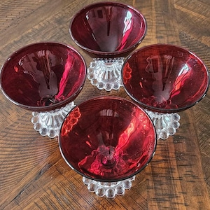 May include: Four vintage ruby red glass dessert bowls. Each bowl has a round, clear glass base with a beaded design. The bowls are a deep red color and sit on a wooden surface.