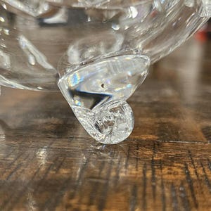 May include: Close-up of a clear, ornate glass bowl with a decorative foot. The bowl has a cut crystal design, reflecting light. The foot of the bowl has a curled, scroll-like shape. The bowl sits on a wooden surface.