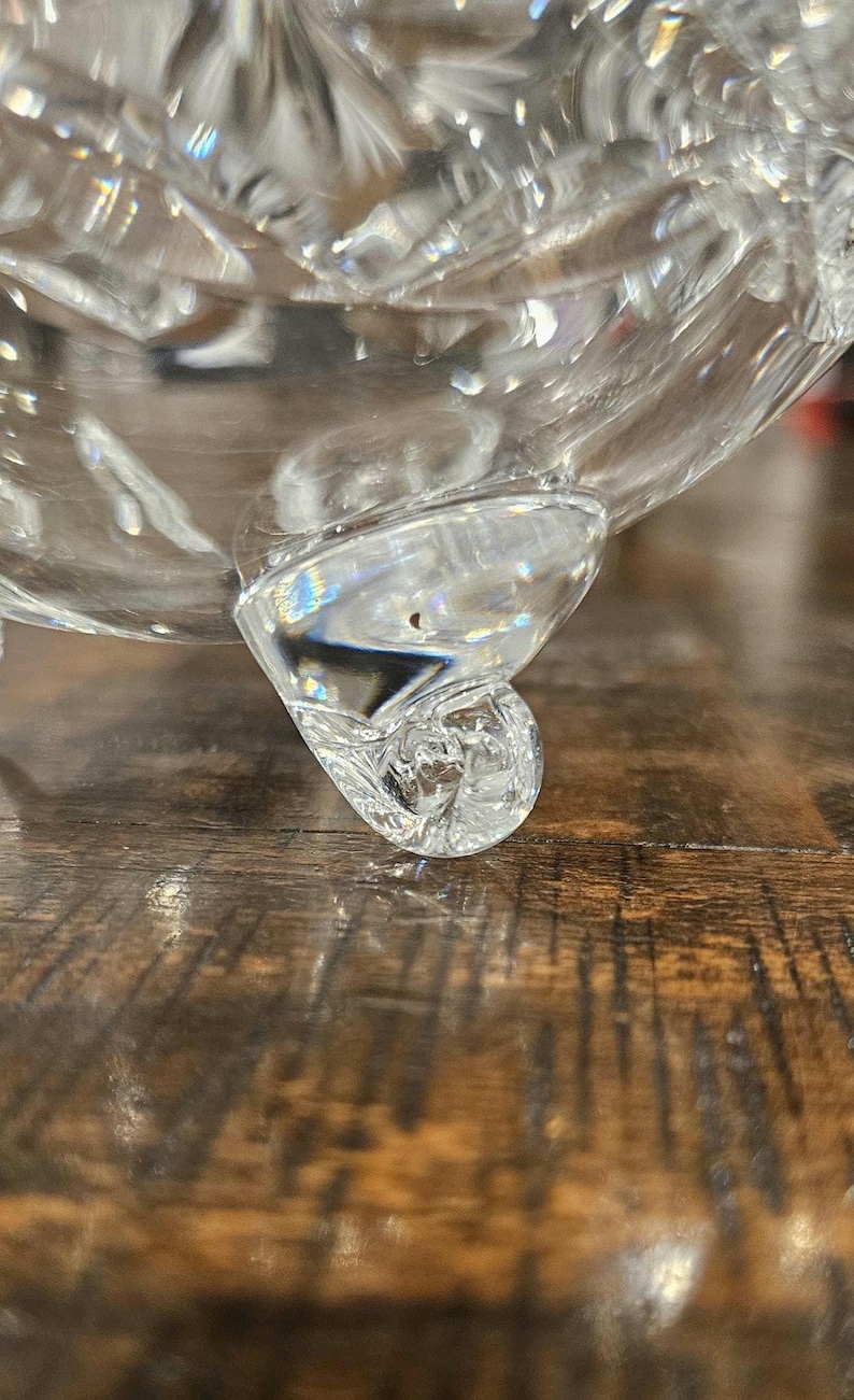 May include: Close-up of a clear, ornate glass bowl with a decorative foot. The bowl has a cut crystal design, reflecting light. The foot of the bowl has a curled, scroll-like shape. The bowl sits on a wooden surface.