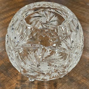 May include: A clear, round crystal bowl with etched floral designs. The bowl has a wide opening and sits on three small feet. The intricate patterns and cuts create a sparkling effect, reflecting light. The bowl is likely used for decorative purposes.