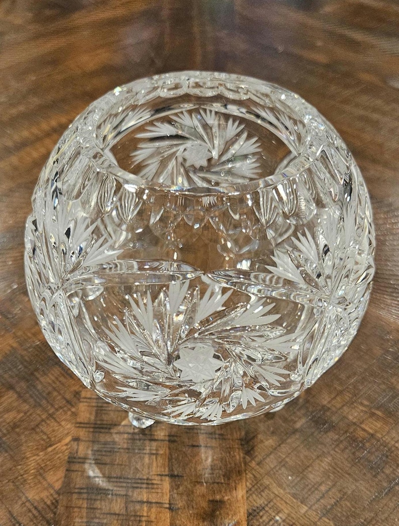 May include: A clear, round crystal bowl with etched floral designs. The bowl has a wide opening and sits on three small feet. The intricate patterns and cuts create a sparkling effect, reflecting light. The bowl is likely used for decorative purposes.