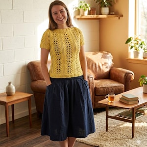 May include: A woman in a yellow, short-sleeved knitted top with an openwork design, paired with a navy blue skirt. The image shows a living room setting with a leather armchair, wooden furniture, and a brick wall.