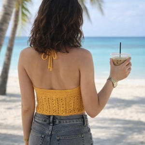 May include: A yellow crochet halter top with a textured design. The top is paired with denim shorts. The background features a beach scene with palm trees and the ocean. A drink with a straw is held in the hand.