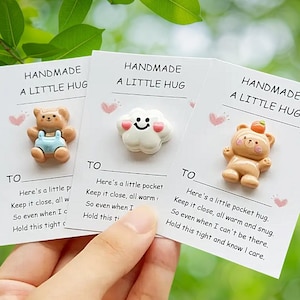 May include: Three handmade cards with small, decorative items. One card features a bear in blue overalls, another a smiling cloud, and the last a bear with a small orange on its head. Each card reads "HANDMADE A LITTLE HUG."