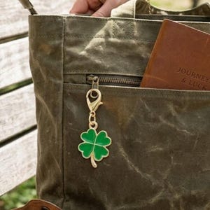 May include: A green four-leaf clover charm with gold-colored trim and a clasp hangs from the zipper of a canvas tote bag. A brown leather journal with the words "JOURNEY & LUCK" is in the bag's pocket.