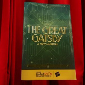 May include: A rectangular program for The Great Gatsby A New Musical featuring a dark green cover with gold geometric art deco lines and text. The bottom section is a yellow horizontal band with the text 2025-2026 KeyBank BROADWAY SERIES.