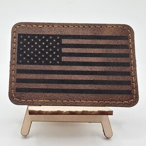 May include: A brown leather patch featuring the American flag design. The flag has black stripes and stars, with a stitched border. The patch is displayed on a small wooden easel.