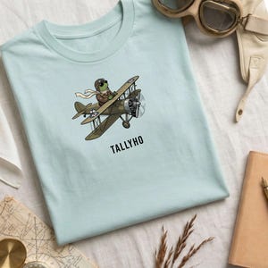 May include: Light blue t-shirt featuring a cartoon frog in a pilot's outfit, flying a biplane. The biplane is olive green with white stars. The word "TALLYHO" is printed below the plane. Accessories include aviator goggles and a leather cap.
