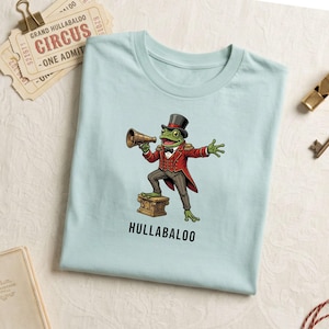 Op de afbeelding: Lichtblauw T-shirt met een cartoon kikker in een circusdirecteur outfit, die een megafoon vasthoudt. De kikker staat op een kleine kist, met het woord "HULLABALOO" eronder. Circustickets en een fluitje zijn zichtbaar.