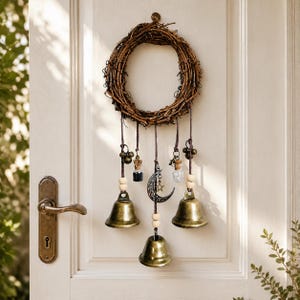 May include: A circular wreath made of brown intertwined twigs hangs on a door. It features five dangling cords adorned with light-colored wooden beads, two small glass vials, a silver-toned crescent moon charm, and three brass-colored bells.