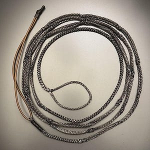 May include: A long, coiled black and white braided rope with a textured surface. One end features a small loop, while the other end transitions into two smooth, brown, flexible strands secured by a black heat-shrink tubing connector.