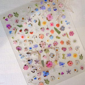 May include: A clear sheet covered with a variety of colorful, miniature flower stickers. The stickers feature a range of colors including pink, blue, yellow, and white, with detailed designs. Some stickers have a 3D effect.