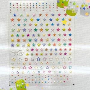 May include: Clear sheet of nail art stickers featuring colorful star and dot designs. The stickers include various shades of pink, orange, yellow, green, blue, and purple. The sheet also has the text "Designed by AR".