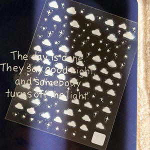 May include: A clear sticker sheet featuring white cloud, star, and heart icons. The text on the sheet reads: The day is done. They say good night, and somebody turns off the light. The stickers are arranged in a repeating pattern on a transparent backing.