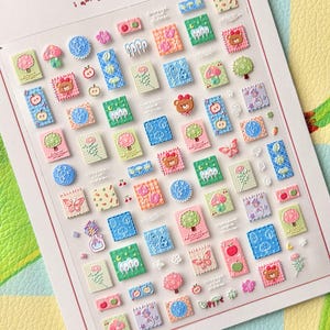 May include: A sheet of colorful, textured stickers with various designs, including apples, bears, trees, and butterflies. The stickers are arranged on a white background with the text "i am a happy apple" at the top. The stickers are in shades of pink, green, blue, and orange.
