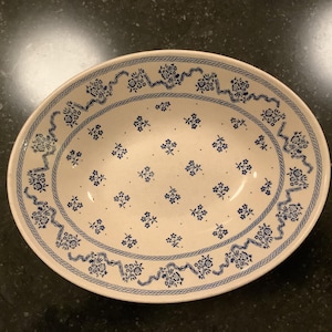 May include: Oval serving dish with a cream-colored base and a blue floral pattern. The rim features a decorative blue border with floral and ribbon motifs. The interior is adorned with small blue floral clusters, creating a classic, elegant aesthetic.