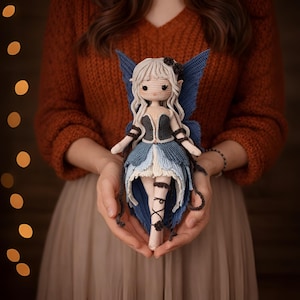 Puede incluir: Una muñeca de hada de ganchillo hecha a mano con cabello gris claro, alas azules y un vestido azul y blanco. La muñeca tiene un corpiño negro y se sostiene en manos abiertas. La muñeca de hada es un artículo único y artesanal.
