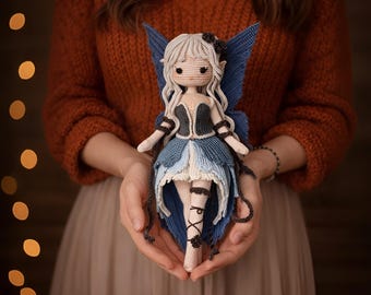 May include: A handmade crochet fairy doll with light gray hair, blue wings, and a blue and white dress. The doll has a black bodice and is held in open hands. The fairy doll is a unique, handcrafted item.