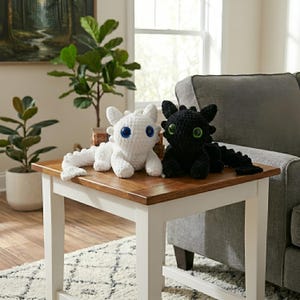 Może przedstawiać: Dwie szydełkowe pluszowe smoki, jeden biały z niebieskimi oczami, a drugi czarny z zielonymi oczami, siedzą na drewnianym stole. Zabawki mają szczegółowe cechy i są umieszczone w pobliżu szarej sofy i roślin.