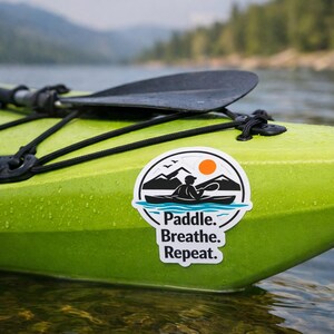 Op de afbeelding: Een limoengroene kajak met een zwarte peddel en een sticker met de tekst "Paddle. Breathe. Repeat." De sticker toont een kajakker, bergen, zon en vogels. De kajak ligt op het water.