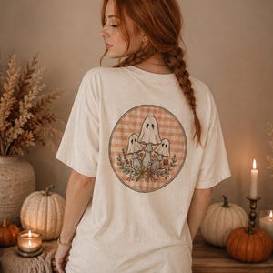 Può includere: Una t-shirt a maniche corte color bianco sporco con una grafica circolare sul retro. Il design presenta tre funghi a forma di fantasma con volti, circondati da fiori e fogliame colorati su uno sfondo a quadretti vichy arancione e bianco.