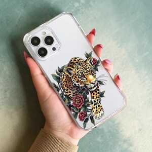 Op de afbeelding: Transparante telefoonhoes met een illustratie van een luipaard. De luipaard heeft een gouden gezicht en zwarte vlekken, met rode rozen en groene bladeren. De hoes wordt vastgehouden in een hand met rode nagellak.