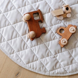 Pode incluir: Três veículos de brinquedo de madeira sobre um tapete circular acolchoado bege claro. O conjunto inclui um camião de madeira castanho escuro, um camião de madeira mais pequeno castanho claro e um carro de madeira castanho escuro.