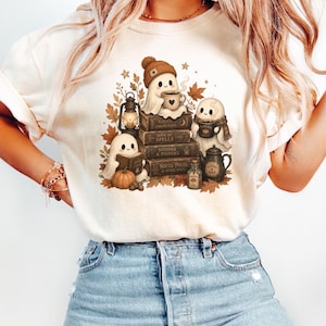 May include: A cream-colored t-shirt featuring a graphic print of three ghosts sitting among stacked books, autumn leaves, a lantern, a pumpkin, and a teapot. The ghosts are wearing hats and scarves, holding mugs, and reading books in a cozy fall scene.