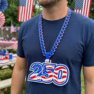 May include: A patriotic necklace with a large pendant displaying the number "250" in red, white, and blue. The chain is blue, white, and red. The necklace is worn over a blue t-shirt, with American flags in the background.