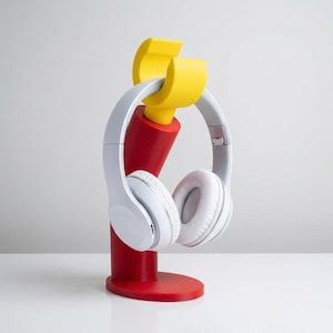 May include: A white pair of headphones rests on a red and yellow headphone stand. The stand has a cylindrical red base and a yellow top designed to hold the headphones. The headphones are white with a padded headband and earcups.