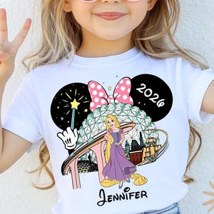 May include: A white t-shirt featuring a colorful graphic of a cartoon character, a pink bow, and the text "2026". The shirt also includes the name "Jennifer" in a script font. The girl is wearing pink sunglasses and making a peace sign.