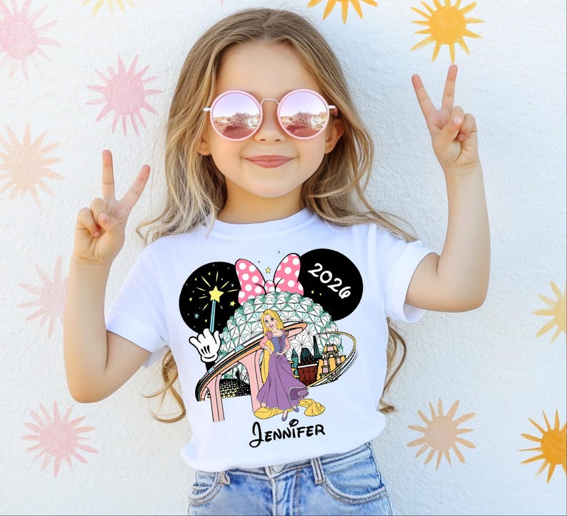 May include: A white t-shirt featuring a colorful graphic of a cartoon character, a pink bow, and the text "2026". The shirt also includes the name "Jennifer" in a script font. The girl is wearing pink sunglasses and making a peace sign.