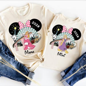 May include: Two cream-colored t-shirts with matching graphics featuring a princess, a Minnie Mouse bow, and the text "2026." One shirt says "Mama," and the other says "Mimi." The shirts are paired with blue denim jeans.