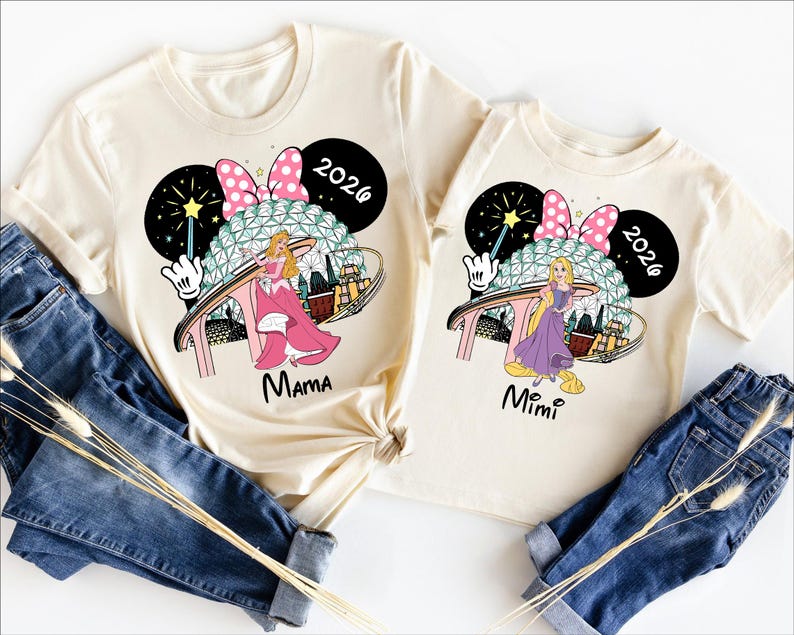 May include: Two cream-colored t-shirts with matching graphics featuring a princess, a Minnie Mouse bow, and the text "2026." One shirt says "Mama," and the other says "Mimi." The shirts are paired with blue denim jeans.