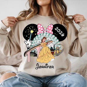 May include: Beige sweatshirt featuring a graphic of Belle from Beauty and the Beast in a yellow gown, with a Minnie Mouse bow, the year 2026, and the name "Jennifer". The design includes a stylized depiction of a theme park.