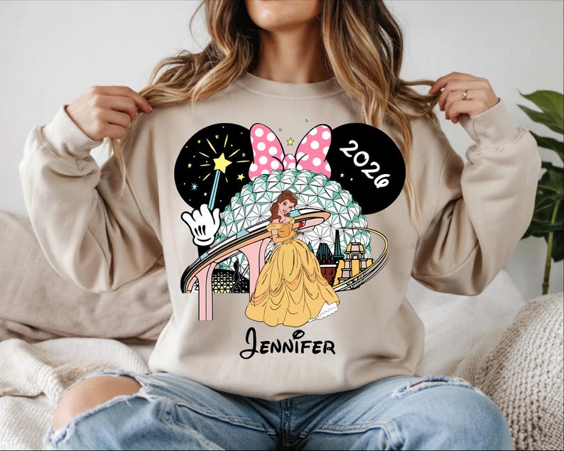 May include: Beige sweatshirt featuring a graphic of Belle from Beauty and the Beast in a yellow gown, with a Minnie Mouse bow, the year 2026, and the name "Jennifer". The design includes a stylized depiction of a theme park.