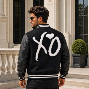 May include: A black varsity jacket featuring long black leather sleeves and a black wool-blend body. The back displays a large, white, textured chenille patch with the letters XO and a heart symbol. It has a striped ribbed collar and cuffs.