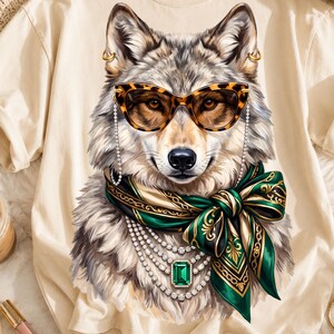 Puede incluir: Una camiseta beige con una ilustración gráfica de un lobo que lleva gafas de ojo de gato con estampado de leopardo y cadena, un pañuelo de seda verde y dorado atado en lazo y varios collares de perlas superpuestos con una gema esmeralda.