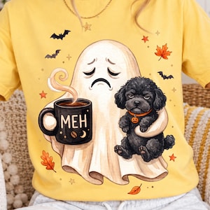 Może przedstawiać: Żółty t-shirt z grafiką smutnego ducha trzymającego czarny kubek z napisem MEH i ikoną ziarna kawy. Obok ducha znajduje się czarny pies z kręconą sierścią, mający na sobie pomarańczową obrożę z zawieszką w kształcie dyni, otoczony nietoperzami.