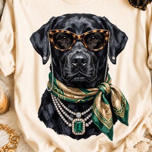 May include: A cream-colored t-shirt featuring a graphic of a black Labrador dog wearing leopard-print cat-eye glasses with a chain, a green and gold patterned silk scarf, and a multi-strand diamond necklace with a large emerald-cut green stone.